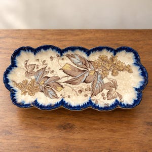 May include: A rectangular ceramic serving tray with a scalloped edge featuring a deep blue border. The center displays a botanical illustration of brown and blue leaves, small berries, and clusters of golden flowers on a cream background.