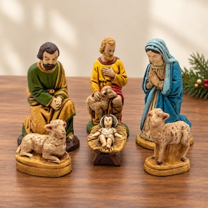 May include: A nativity scene set featuring five hand-painted figurines: a kneeling figure in green and gold, a figure in yellow and red with a sheep, a figure in blue and tan, a baby in a manger, and two separate sheep figurines.