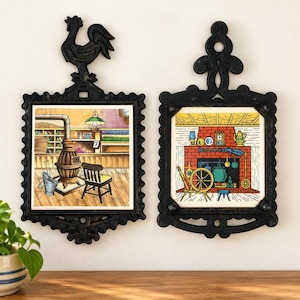 May include: Two black cast iron wall plaques featuring square ceramic tiles with colorful illustrations. The left tile depicts a wood stove in a room, while the right tile shows a fireplace with a spinning wheel and a rifle mounted above the mantle.