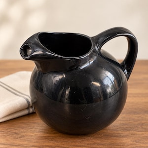May include: A glossy black ceramic pitcher with a rounded, spherical body, a wide open top, and a curved handle. The pitcher features a prominent, short pouring spout extending from the side, with a smooth, reflective surface finish throughout.