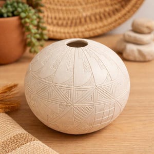 May include: A cream-colored, spherical vase with a small opening at the top. The surface is intricately carved with geometric patterns, including triangles and lines. The vase is displayed on a wooden surface.