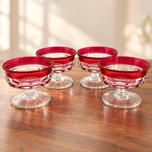 May include: A set of four glass dessert bowls featuring a deep red rim and a clear, textured base with a geometric pattern. Each bowl stands on a short, sturdy pedestal stem and a circular foot, showcasing a clear glass bottom and a vibrant red top.