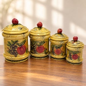 May include: A set of four graduated ceramic canisters in a yellow-gold glaze, each featuring a raised relief design of grapes and apples. Every canister includes a matching lid topped with a small, glossy red apple-shaped handle.