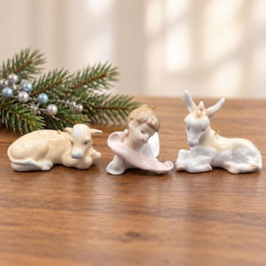 May include: Three porcelain ornaments featuring a reclining calf in cream, a child figure holding a light pink ribbon, and a reclining donkey in cream and white. Each ornament includes a small gold loop for hanging on a tree.