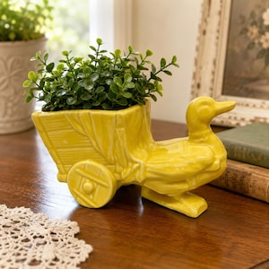 May include: A bright yellow ceramic planter shaped like a duck pulling a small cart. The duck has a smooth, glossy finish and is filled with a lush, green artificial plant with small, rounded leaves. The cart features molded wheel details.