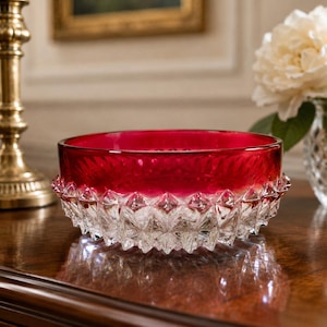 May include: A ruby red glass bowl with a clear, diamond-cut base. The bowl has a smooth, glossy interior and a textured, faceted exterior. The decorative bowl is likely used for serving or display.