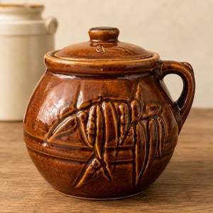 May include: A round, brown glazed ceramic pot with a matching lid and a single side handle. The exterior features a raised, embossed relief pattern depicting stylized plant leaves and stems wrapping around the body of the vessel.