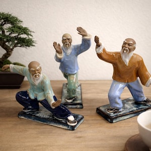 May include: Three ceramic figurines of elderly men in martial arts poses. Each figure is dressed in different colored clothing: light blue, blue, and orange. They stand on rectangular bases with a textured, dark blue finish. A bonsai tree is visible in the background.