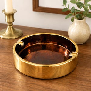 May include: A round, low-profile ashtray featuring a polished gold-tone exterior and a dark, metallic copper-colored interior basin. The design includes two symmetrical notches on the rim for resting items, with a smooth, reflective finish.