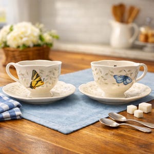 May include: Two white teacups and saucers with butterfly designs. One cup features a yellow butterfly, the other a blue butterfly. Both cups have a scalloped rim and sit on a light blue placemat. Two silver spoons and sugar cubes are also present.