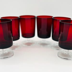 May include: A set of six matching drinking glasses with deep red bowls and clear glass stems and circular bases. Each glass features a smooth, cylindrical shape that tapers slightly toward the bottom, resting on a sturdy, transparent pedestal foot.
