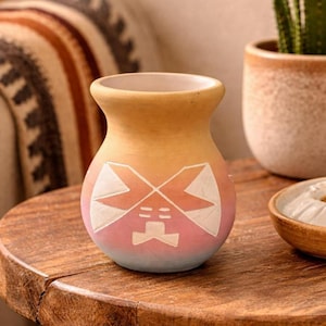 May include: A small, hand-painted ceramic vase with a unique design. The vase features a gradient of colors, transitioning from a light yellow at the top to a soft pink and blue at the base. White geometric shapes are painted on the front.