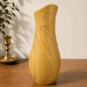 May include: A tall, mustard-yellow ceramic vase with a matte finish. The vase features a slender, elongated neck that widens toward the base, decorated with subtle, swirling, raised vertical ridges that wrap around the body in a continuous pattern.