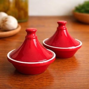 May include: Two red ceramic tagine pots with white trim. The tagines have conical lids and rounded bases. They are displayed on a wooden surface, with other kitchen items in the background.