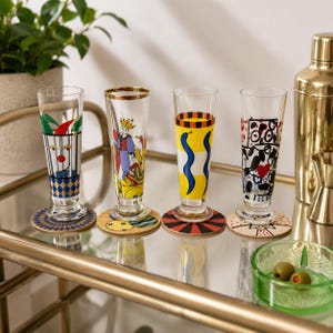 May include: Four tall, clear glass tumblers with distinct colorful designs, including a jester, a royal figure, blue snakes on a yellow background, and abstract black and red patterns. Each glass rests on a circular coaster with matching motifs.