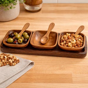 May include: A wooden serving tray with three square bowls and small wooden spoons. The bowls contain olives, nuts, and are empty. The tray is dark brown, and the bowls are a lighter wood color. A linen napkin and a pile of nuts are in the foreground.