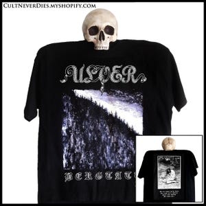 Könnte beinhalten: Schwarzes T-Shirt mit dem Bandnamen "ULVER" in einer dekorativen Schriftart über einer Berglandschaft. Das Wort "BERGTAKT" befindet sich unter dem Bild. Auf der Rückseite des T-Shirts befindet sich ein kleines Bild einer Person. Ein Schädel liegt auf dem T-Shirt.