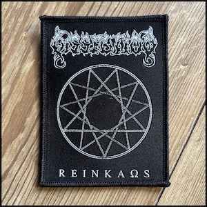 May include: Black rectangular patch with white embroidered text and design. The top text reads "Dissection" in a stylized font. Below is a white geometric star pattern within a circle. The bottom text reads "REINKAOS". The patch has a black border.