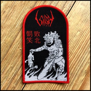 May include: Black patch with a red border featuring a white and black illustration of a demon-like figure holding a smaller figure. The patch has red text at the top and in a vertical column. The patch is on a wooden surface.