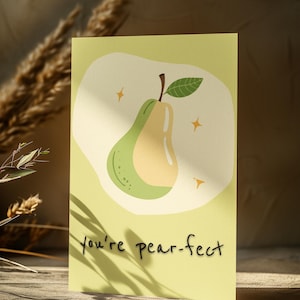 May include: A light green greeting card with a pear illustration in green and beige, with a brown stem and green leaf. The words "you're pear-fect" are written in black script. The card is on a wooden surface.