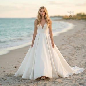 May include: A floor-length ivory gown featuring a deep V-neckline, thin shoulder straps, and a fitted bodice that transitions into a full, pleated A-line skirt with a train. The dress has a smooth, solid finish and a clean, structured silhouette.