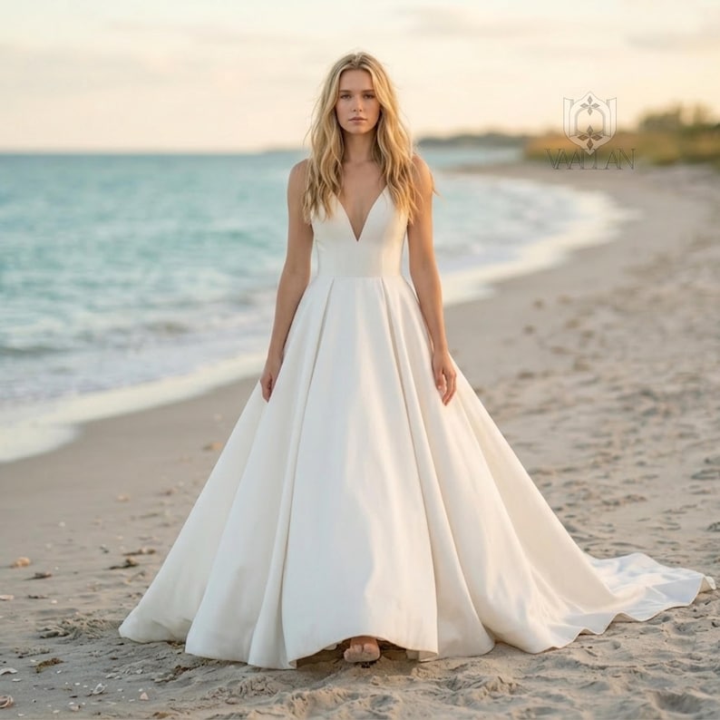 May include: A floor-length ivory gown featuring a deep V-neckline, thin shoulder straps, and a fitted bodice that transitions into a full, pleated A-line skirt with a train. The dress has a smooth, solid finish and a clean, structured silhouette.