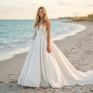 May include: A floor-length ivory white gown featuring a deep V-neckline, thin shoulder straps, and a fitted bodice that transitions into a voluminous, pleated A-line skirt with a long train, crafted from a smooth, solid-colored fabric.