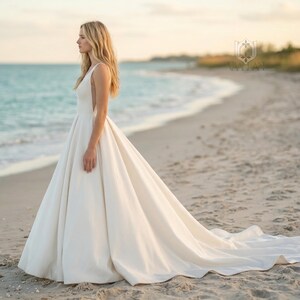 May include: A floor-length, sleeveless ivory wedding dress with a fitted bodice, a deep V-neckline, and a full, voluminous skirt that extends into a train. The dress features a clean, solid fabric design with no visible embellishments or patterns.