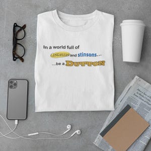 Könnte beinhalten: Weißes T-Shirt mit dem Text "In a world full of Costanzas and stinsons...be a DUTTON" in blauen und gelben Buchstaben. Das Shirt liegt auf einer grauen Oberfläche mit Smartphone, Brille, Kopfhörern, Kaffeebecher und Zeitung.