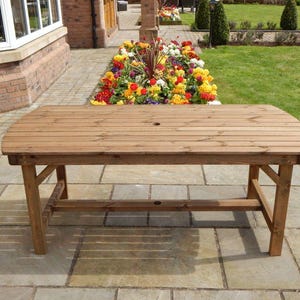May include: A wooden garden table with a slatted top and curved ends. The table is approximately 152 cm long and 76 cm wide, with a central hole. It has four sturdy legs and is placed on a stone patio, ideal for outdoor dining.