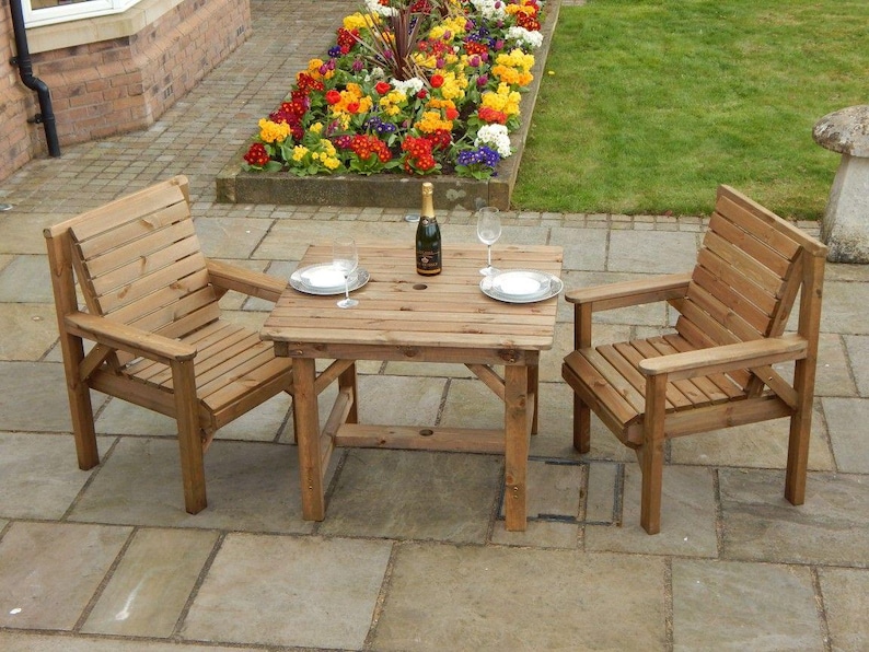 Op de afbeelding: Een houten terrasmeubelset met twee fauteuils en een vierkante tafel. De stoelen en tafel zijn gemaakt van lichtgekleurd hout. Een fles champagne en twee wijnglazen staan op de tafel. De set staat op een stenen terras.