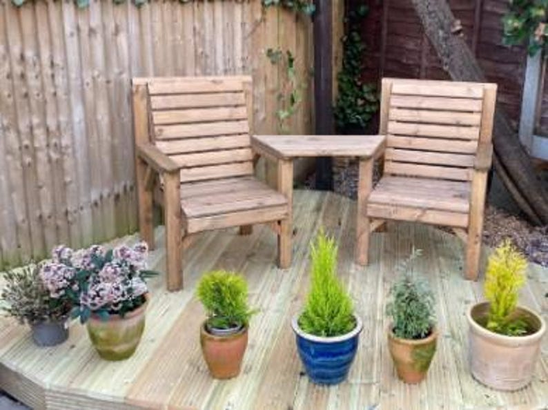 Peut inclure: Ensemble de mobilier de jardin en bois comprenant deux chaises et une petite table. Les chaises et la table sont en bois clair. Diverses plantes en pot, dont des plantes &agrave; fleurs et de petits conif&egrave;res, sont dispos&eacute;es sur la terrasse en bois.