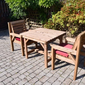 Op de afbeelding: Een houten eethoek voor buiten met een vierkante tafel en twee stoelen. De tafel en stoelen zijn gemaakt van lichtgekleurd hout. De stoelen hebben rode kussens. De set staat op een stenen terras, omringd door groen.