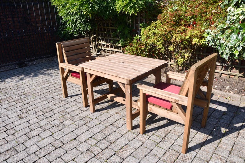 Op de afbeelding: Een houten eethoek voor buiten met een vierkante tafel en twee stoelen. De tafel en stoelen zijn gemaakt van lichtgekleurd hout. De stoelen hebben rode kussens. De set staat op een stenen terras, omringd door groen.