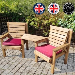 Peut inclure: Deux chaises de jardin en bois avec une petite table attach&eacute;e. Les chaises ont une finition en bois naturel et des coussins de si&egrave;ge rouges. Les chaises sont pos&eacute;es sur un patio pav&eacute;, avec de la verdure en arri&egrave;re-plan.