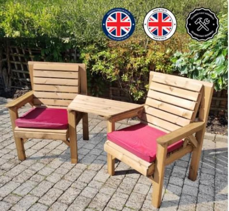 Peut inclure: Deux chaises de jardin en bois avec une petite table attach&eacute;e. Les chaises ont une finition en bois naturel et des coussins de si&egrave;ge rouges. Les chaises sont pos&eacute;es sur un patio pav&eacute;, avec de la verdure en arri&egrave;re-plan.