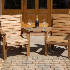 Peut inclure: Deux chaises de jardin en bois et une petite table. Les chaises ont des dossiers &agrave; lattes et des accoudoirs. Une bouteille de champagne et deux verres &agrave; vin sont sur la table. Le mobilier est en bois clair.