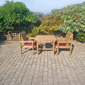 Op de afbeelding: Een houten terrasmeubelset met een kleine vierkante tafel en twee stoelen. De stoelen hebben rode kussens. De set is gemaakt van lichtgekleurd hout en staat op een stenen terras. Groene bladeren zijn op de achtergrond.