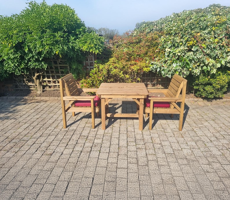 Op de afbeelding: Een houten terrasmeubelset met een kleine vierkante tafel en twee stoelen. De stoelen hebben rode kussens. De set is gemaakt van lichtgekleurd hout en staat op een stenen terras. Groene bladeren zijn op de achtergrond.