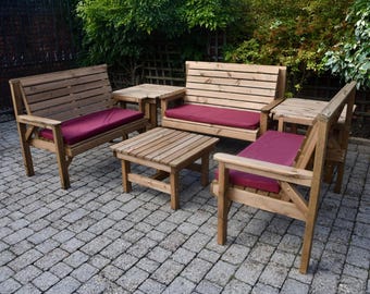 May include: Outdoor wooden furniture set with a natural finish. The set includes two armchairs, a loveseat, and two small square tables. Each seat has a burgundy cushion. The furniture is arranged on a brick patio, surrounded by greenery.