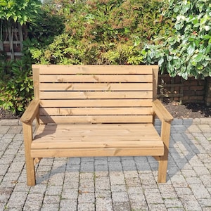May include: A light brown wooden bench with a slatted back and seat, and armrests. The bench is outdoors, set on a brick patio, with lush green foliage in the background. The bench is approximately 122 cm wide.