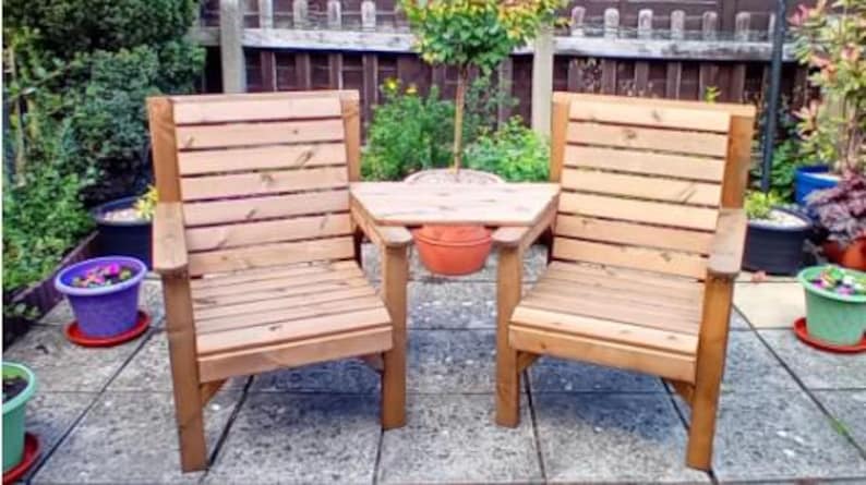 Peut inclure: Deux chaises de jardin en bois avec une petite table centrale. Les chaises et la table sont en bois brun clair et pr&eacute;sentent un design &agrave; lattes. Les chaises ont des accoudoirs. La sc&egrave;ne se d&eacute;roule sur un patio avec des plantes en pot en arri&egrave;re-plan.