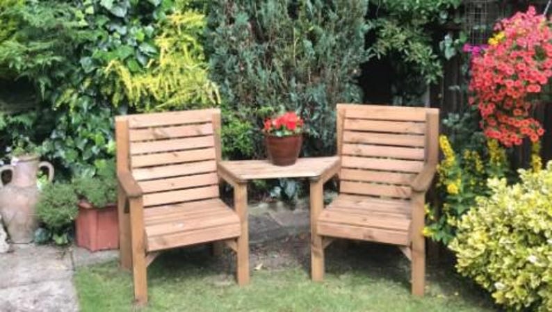 Peut inclure: Deux chaises de jardin en bois et une petite table. Les chaises ont des dossiers &agrave; lattes et des accoudoirs. Un petit pot de fleurs marron est pos&eacute; sur la table. Le mobilier est en bois brun clair, sur de l'herbe.