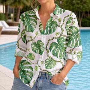 May include: White button-down shirt with a green monstera leaf pattern. The long-sleeved shirt has a V-neck and is paired with blue jeans. The tropical leaf design is repeated across the shirt.