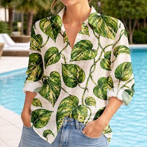 May include: A cream-coloured button-down shirt with a botanical leaf pattern. The long-sleeved shirt features large green and yellow leaves. The sleeves are rolled up, and the shirt is unbuttoned at the top.