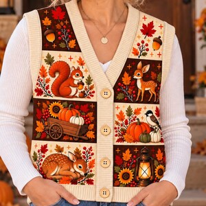 May include: A cream-colored V-neck sweater vest featuring a button-front closure and a multi-panel autumn print. The design includes illustrations of a squirrel, a deer, a rabbit, pumpkins, sunflowers, acorns, fall leaves, and a lantern.