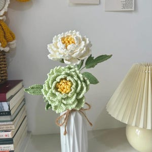 Può includere: Un vaso di ceramica bianca contiene due fiori all'uncinetto: una peonia bianca e una peonia verde. I fiori hanno centri gialli e foglie verdi. Il vaso è legato con spago marrone.