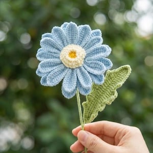 Op de afbeelding: Een gehaakte blauwe madelief met een gele kern en groene stengel en blad. De handgemaakte bloem wordt tegen een vage groene achtergrond gehouden. De bloemblaadjes van de madelief zijn zachtblauw en de kern is getextureerd geel.