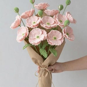 Puede incluir: Un ramo de amapolas rosas de ganchillo con tallos y capullos verdes, envuelto en papel marrón y atado con hilo. Las flores hechas a mano tienen pétalos detallados y son sostenidas por una persona.