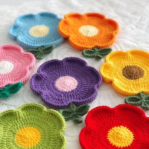 Könnte beinhalten: Eine Sammlung farbenfroher, gehäkelter Untersetzer in Blumenform. Die Untersetzer weisen verschiedene Farben auf, darunter Blau, Orange, Rosa, Lila, Gelb, Grün und Rot, mit kontrastierenden Zentrumsfarben und grünen Blattakzenten. Diese handgefertigten Untersetzer verleihen einen Hauch von Laune.